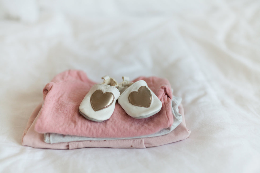 White baby shoes with heart design on folded pink and gray baby clothes on a bed