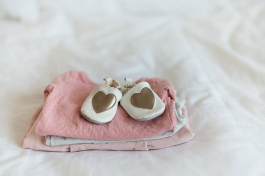 White baby shoes with heart design on folded pink and gray baby clothes on a bed